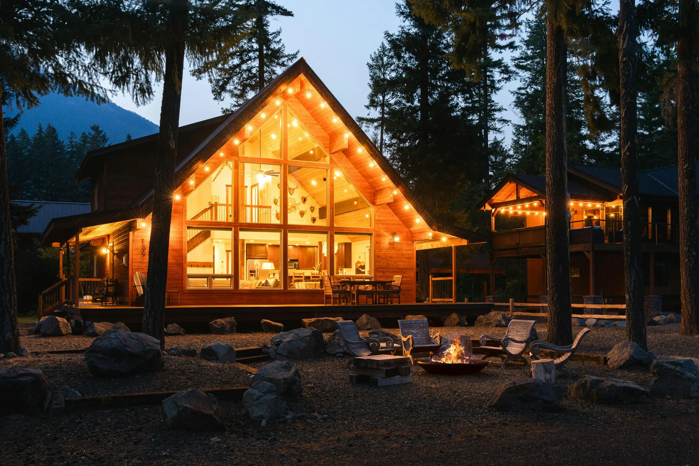 Chalet de madera con fachada de vidrio iluminada al anochecer y una fogata al frente