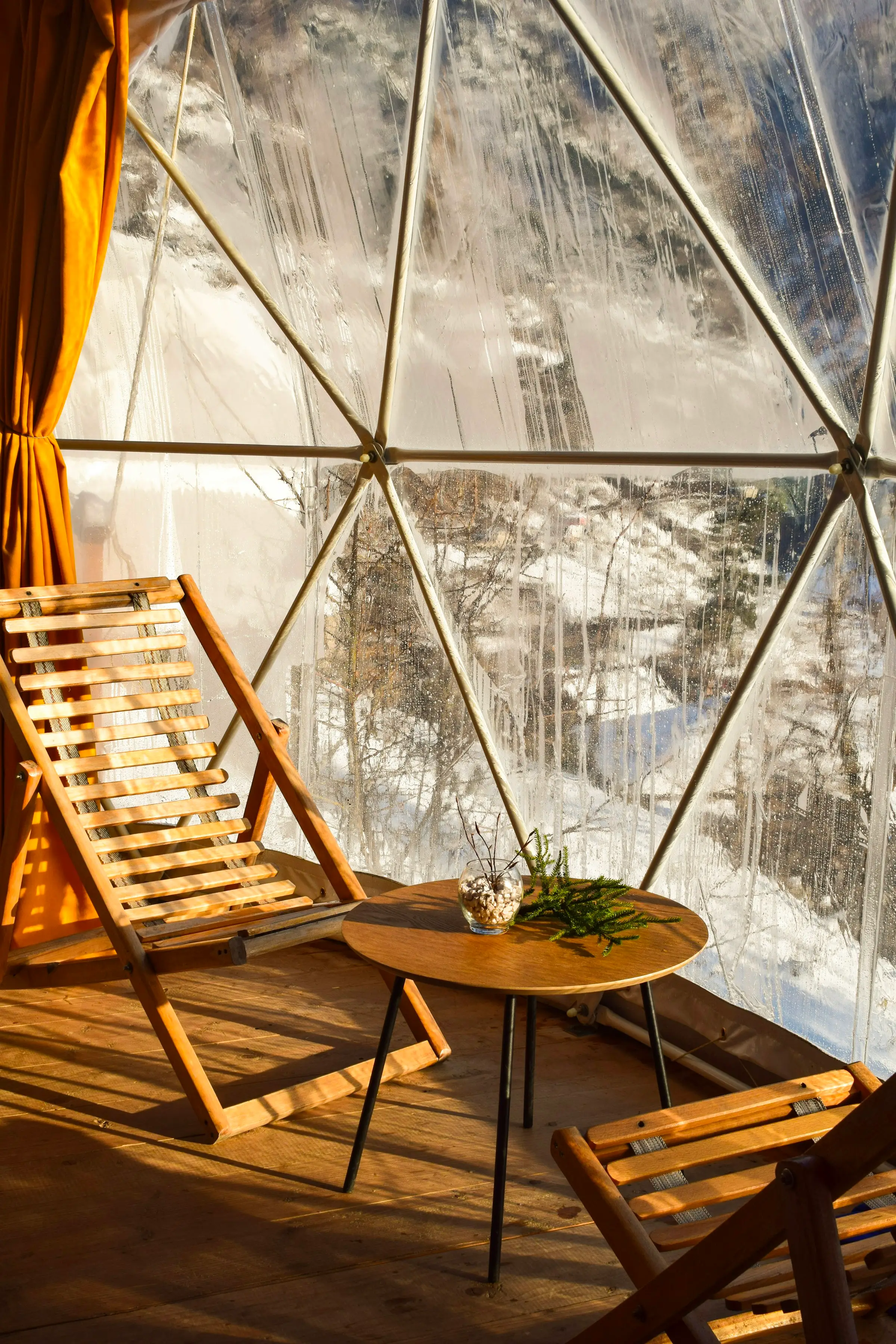 Interior de un domo geodésico transparente con una silla de madera mirando al bosque