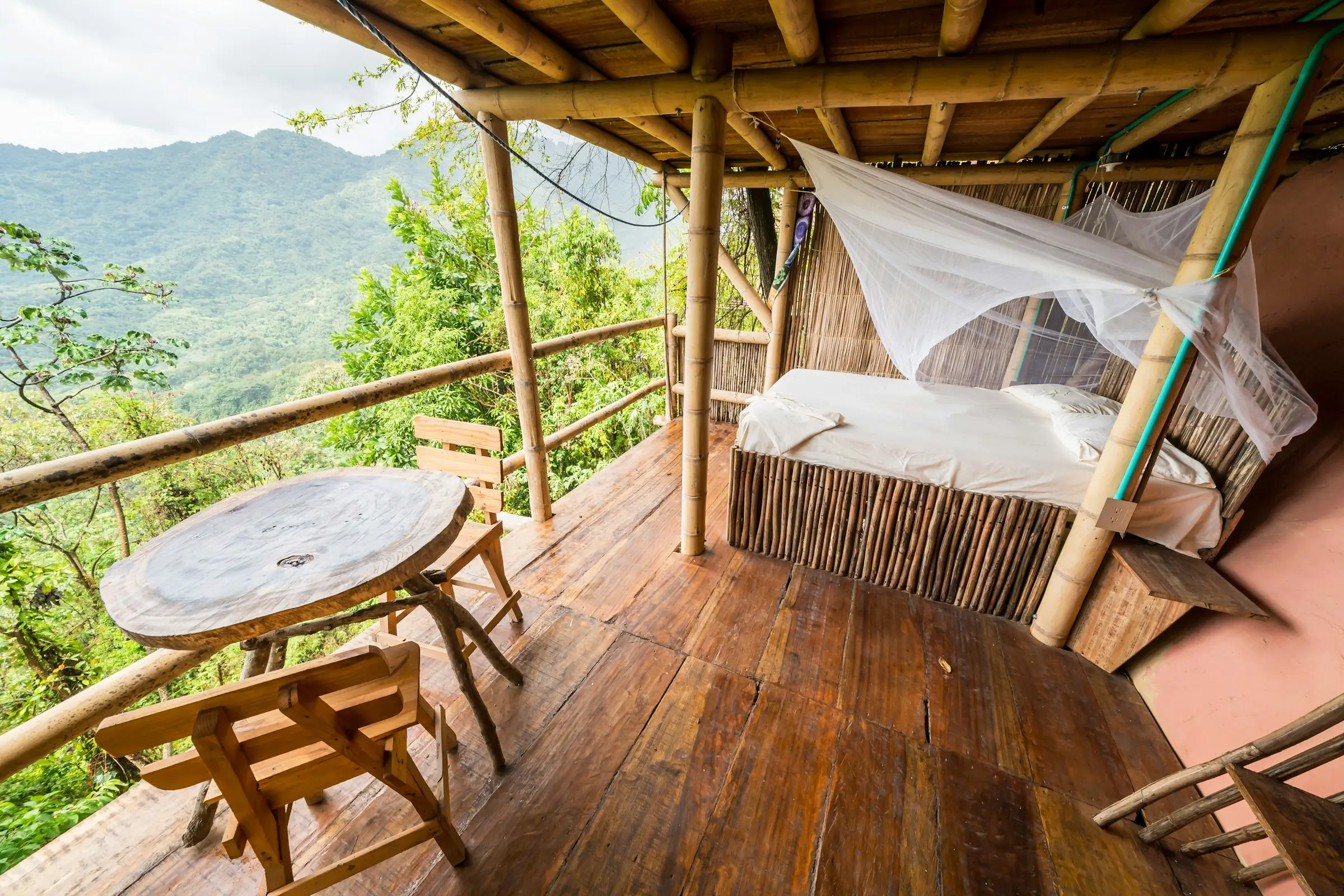 Terraza de bambú con cama con toldillo y una mesa, frente a montañas verdes