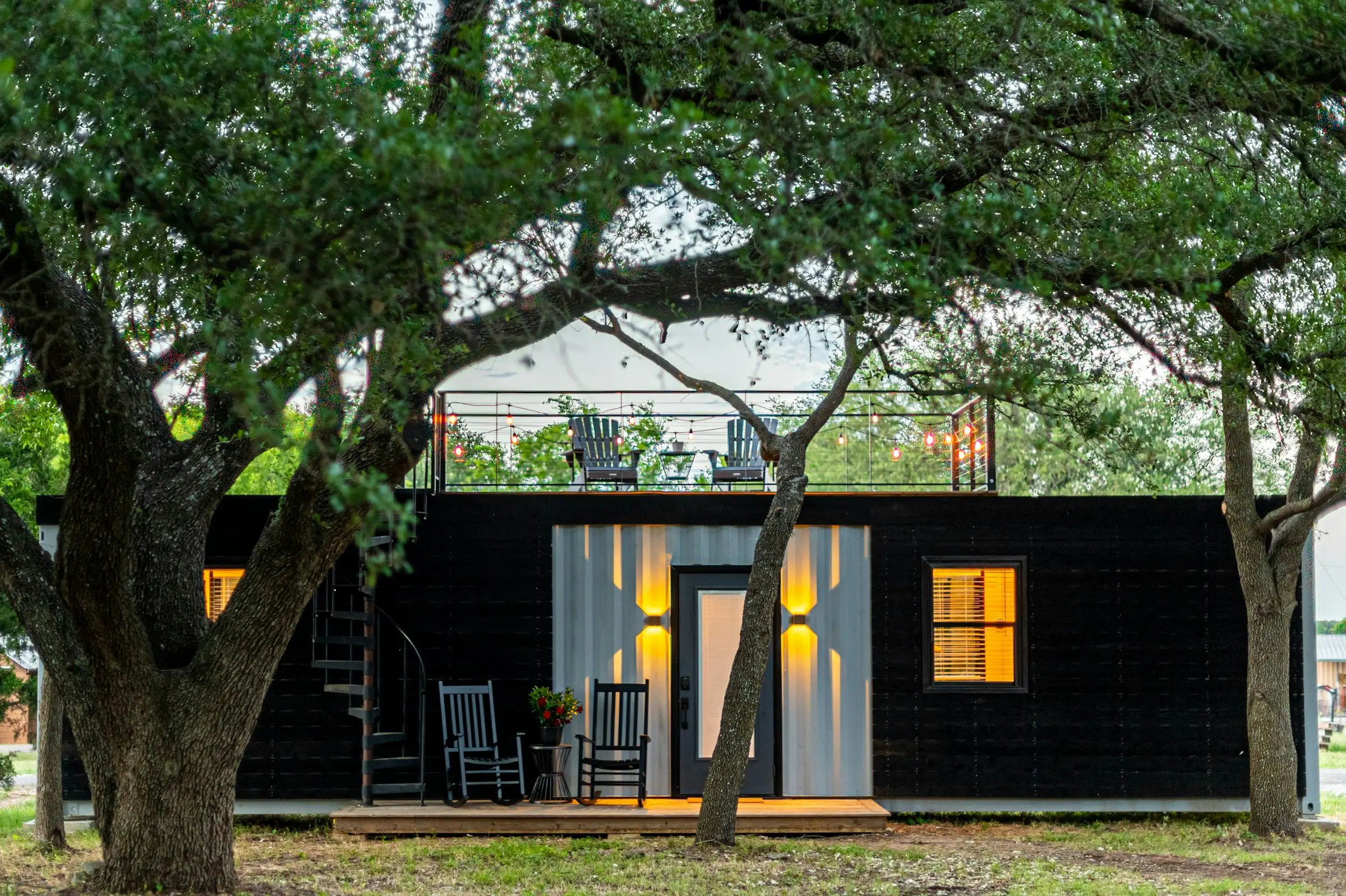 Cabaña pequeña de madera negra con luces encendidas, bajo un árbol grande